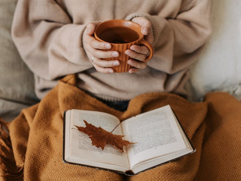 A person in a beige oversized sweater holding a ribbed terracotta mug of tea over an open book with a dried autumn leaf on the pages.