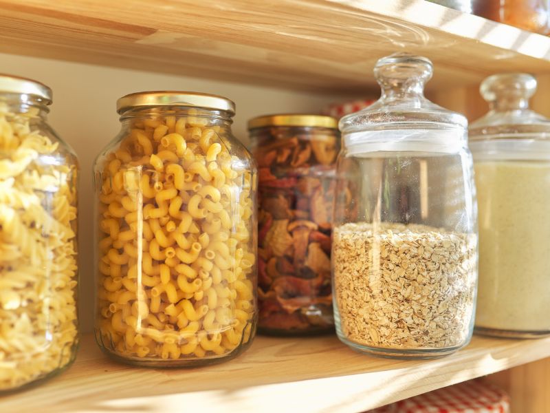 Row of clear glass jars filled with pasta, oats, and dried fruit on a light wooden pantry shelf with warm natural sunlight.