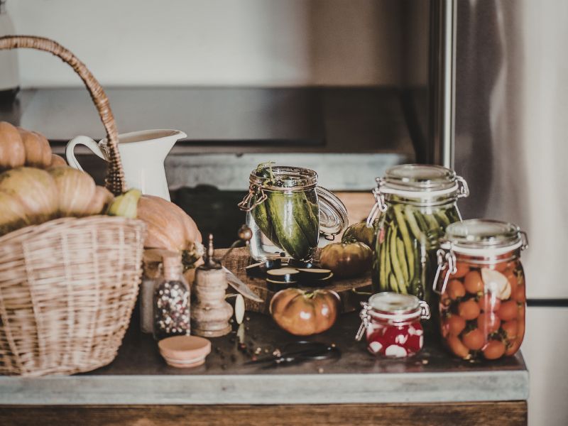 A wooden countertop with a basket of pumpkins, fresh tomatoes, and several glass flip-top jars containing pickled cucumbers, green beans, and cherry tomatoes.